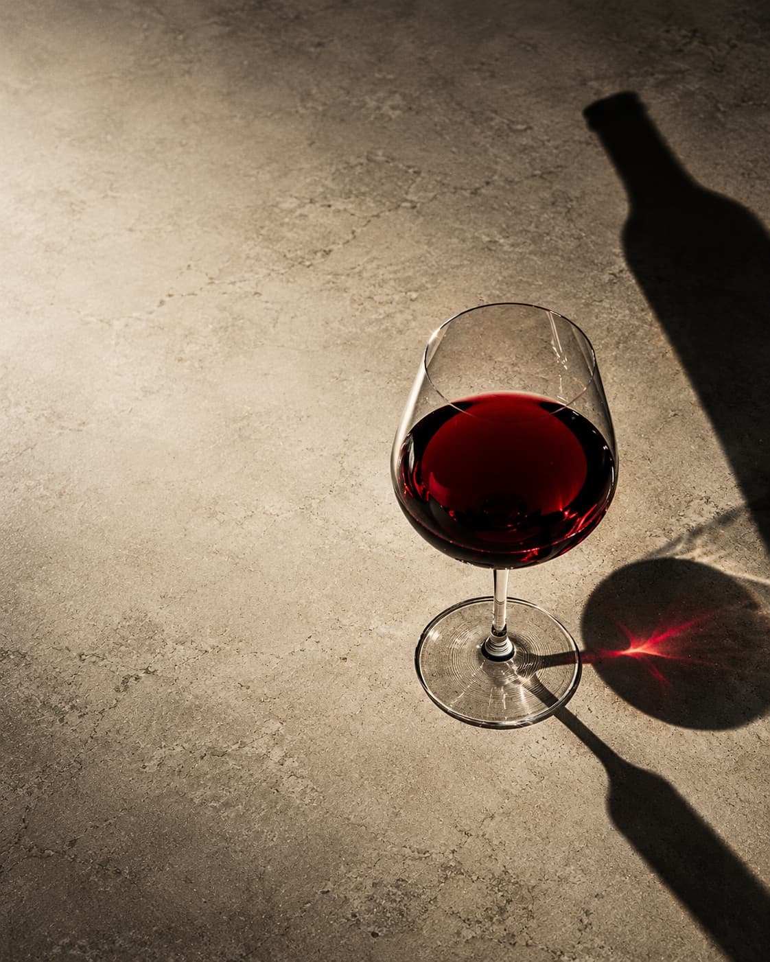 Un verre de vin rouge sur du béton, éclairé par une lumière dure qui projette l'ombre d'une bouteille